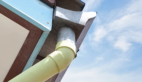 Metal gutter outlet connected to a pale green plastic downspout elbow under a roof eave
